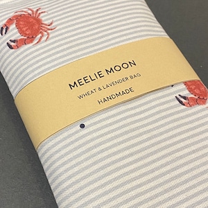 May include: A handmade wheat and lavender bag featuring a blue and white striped design and crab motifs. The bag is wrapped with a brown paper band that reads "MEELIE MOON WHEAT & LAVENDER BAG HANDMADE".