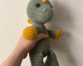 May include: Crocheted dinosaur toy in shades of gray and yellow. The handmade plushie features a textured body, small arms and legs, and a spiky crest. The dinosaur has black button eyes and is approximately 8 inches tall.