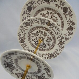 Könnte beinhalten: Ein dreistöckiger Kuchenständer mit einer goldenen Metallbasis und einem blauen Band. Jede Etage ist ein weißer Keramikteller mit einem schwarzen und braunen Blumenmuster.