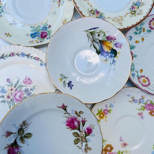 Peut inclure: Une collection de sept soucoupes et assiettes en céramique blanche ornées de divers motifs floraux, notamment des roses, des tulipes et des petites fleurs sauvages. Chaque pièce présente des bords dorés et festonnés.