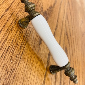 May include: A cabinet handle featuring a smooth, white, cylindrical ceramic center section. The handle is supported by two ornate, weathered brass-toned metal ends with a textured, aged finish and decorative, tiered, spindle-shaped finials.