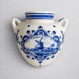 May include: A white ceramic wall pocket vase featuring blue hand-painted floral patterns and a central oval medallion depicting a windmill landscape with the text Holland. The vessel has two side handles and a flat back for wall mounting.