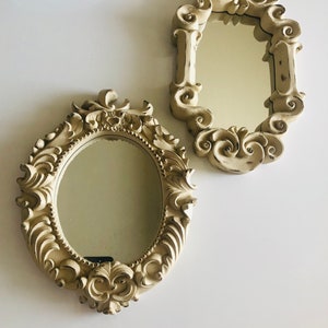 May include: Two ornate oval mirrors with decorative frames. The frames are a light beige color with intricate details. The mirrors are both oval shaped and have a reflective surface.