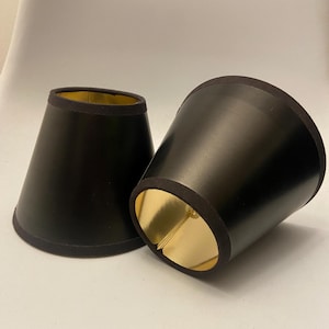 May include: Two conical lamp shades featuring a smooth matte black exterior finish and a reflective metallic gold interior lining. Each shade has a circular opening at the top and bottom with a black fabric trim along the edges for a clean finish.