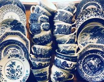 May include: A collection of blue and white porcelain teacups and saucers with various landscape and floral designs. The teacups are stacked, and the plates are arranged around them on a wooden tray. The overall aesthetic is vintage and classic.