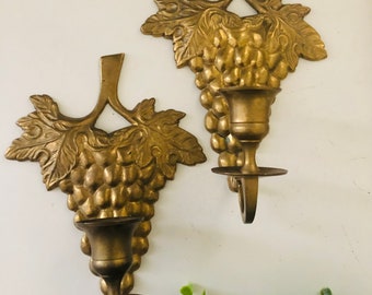 May include: A pair of gold-toned brass wall sconces with a grapevine design. Each sconce has a candle holder and a hook for hanging.