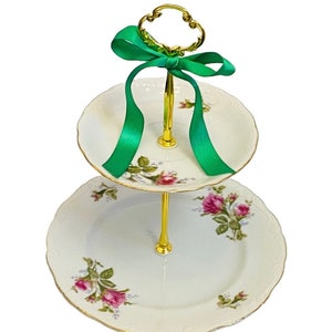 May include: Two-tiered white porcelain cake stand with a gold trim and floral design. The stand has a gold metal handle with a green ribbon.