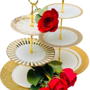 Puede incluir: Un soporte de servicio de cuatro niveles con platos blancos y ribetes dorados, sostenido por una estructura dorada. Decorado con rosas rojas, es perfecto para presentar pasteles o aperitivos.