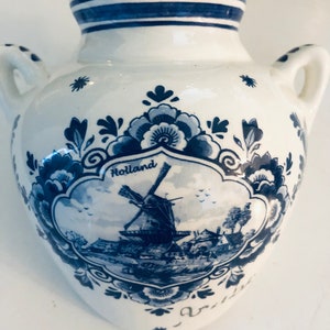 Puede incluir: Un jarrón de cerámica blanca con diseños de Delftware azul y blanco. El jarrón presenta una escena de molino de viento con la palabra "Holland" y un patrón floral.