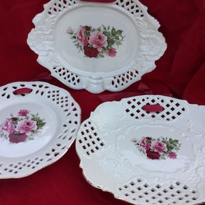 May include: Three white ceramic plates with a lace design and floral patterns. The plates have a raised edge and a gold trim. The flowers are pink and red.