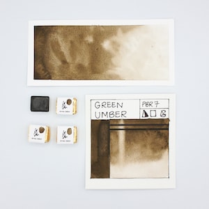 May include: A watercolour paint pan with a brown pigment labelled "Green Umber" and the colour index PBR7. The paint pan is surrounded by three other paint pans, two of which are labelled "Green Umber".