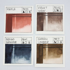 May include: Four watercolor paint swatches, each with a different color and pigment code. The colors are maple, violet gray, indigo genuine, and green umber.
