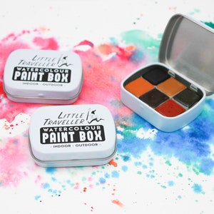 May include: Two white metal tins with the words "Little Traveller Watercolour Paint Box" printed on the front. The tins are open and one shows a set of six watercolor paint pans in shades of orange, brown, and black.