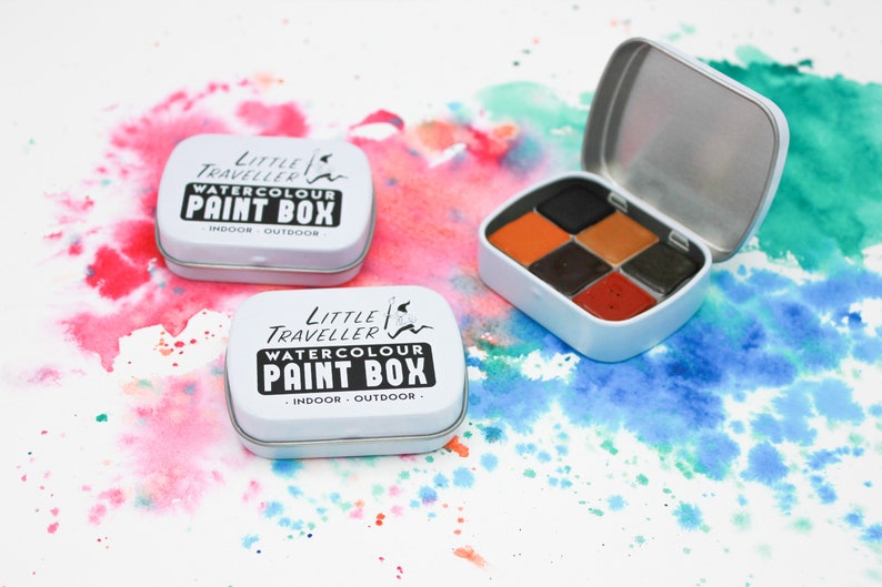 May include: Two white metal tins with the words "Little Traveller Watercolour Paint Box" printed on the front. The tins are open and one shows a set of six watercolor paint pans in shades of orange, brown, and black.