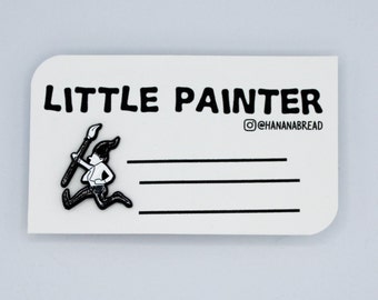 May include: Black and white enamel pin featuring a cartoon character running with a paintbrush. The pin says "LITTLE PAINTER" and "@HANANABREAD" on the card.
