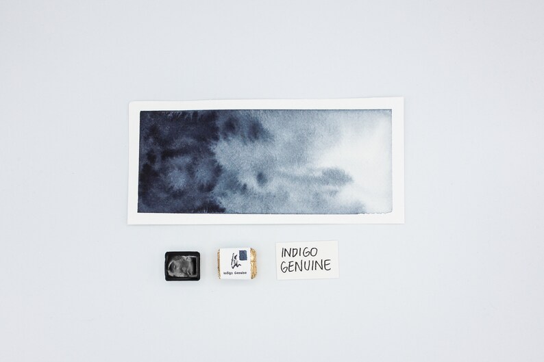 May include: A watercolor wash of indigo blue paint on white paper. A small pan of indigo watercolor paint and a label that reads "INDIGO GENUINE" are below the wash.