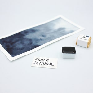 May include: A watercolor paint pan with a dark blue pigment. The pan is labeled "Indigo Genuine" and is next to a watercolor wash sample on a white surface.