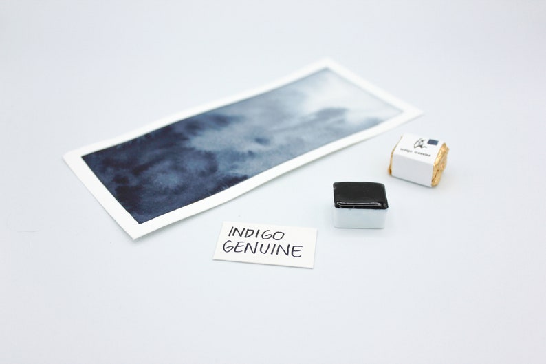 May include: A watercolor paint pan with a dark blue pigment. The pan is labeled "Indigo Genuine" and is next to a watercolor wash sample on a white surface.