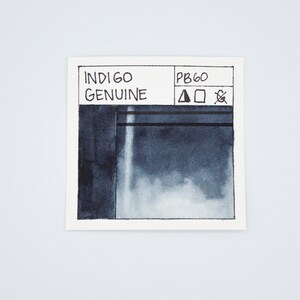 May include: A small, white card with black text and a dark blue watercolor swatch. The text reads "INDIGO GENUINE" and "PB60". There are three symbols next to the PB60: a triangle, a square, and a circle with a diagonal line through it.