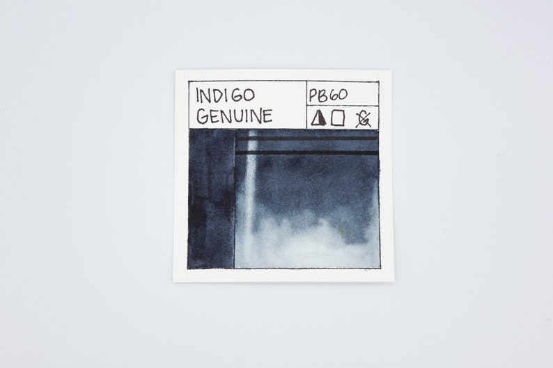 May include: A small, white card with black text and a dark blue watercolor swatch. The text reads "INDIGO GENUINE" and "PB60". There are three symbols next to the PB60: a triangle, a square, and a circle with a diagonal line through it.