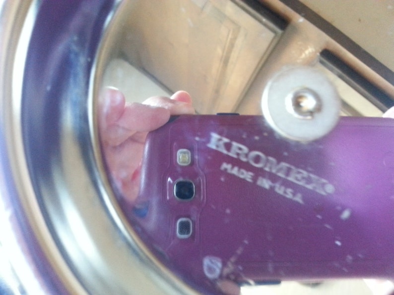 Pu&ograve; includere: Un coperchio metallico circolare Kromex con una superficie riflettente. Il coperchio presenta una manopola centrale e la scritta KROMEX MADE IN USA stampata sulla superficie. La finitura metallica &egrave; liscia e lucida, riflettendo un telefono viola.
