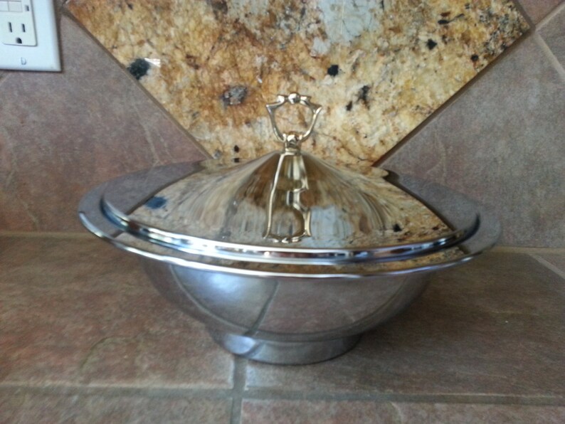 Pu&ograve; includere: Una ciotola da portata in metallo color argento lucido con coperchio a cupola abbinato. Il coperchio presenta una maniglia decorativa centrale con un design ad anello. La ciotola ha un bordo largo e piatto e una base affusolata.