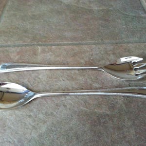 May include: A pair of polished silver-tone metal serving utensils consisting of a large spoon and a matching fork with three tines. Both pieces feature a slender handle with a decorative beaded edge detail along the outer border.