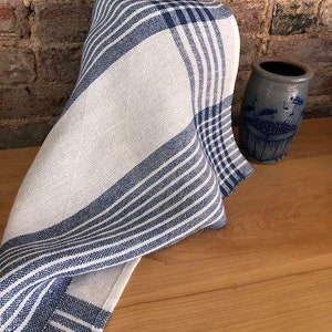 May include: A white and blue striped kitchen towel with a checkered pattern. The towel is folded and draped over a wooden surface.