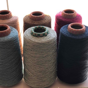 Könnte beinhalten: Sechs Garnspulen in verschiedenen Farben, darunter Blau, Grau, Rosa und Braun. Jede Spule hat eine zylindrische Form mit einem Pappkern und einer strukturierten, hellfarbigen Oberseite. Das Garn scheint eng gewickelt zu sein und ist für Textilprojekte geeignet.