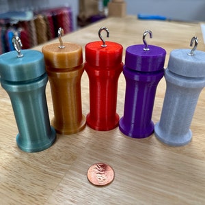 Puede incluir: Cinco devanadores de hilo de diferentes colores con ganchos de metal. Los devanadores son azul verdoso, dorado, rojo, morado y gris.