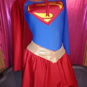 Könnte beinhalten: Ein blaues Langarmshirt mit einem rot-gelben Superman-Logo auf der Brust, kombiniert mit einem roten Rock mit metallisch goldenem Bund und einem passenden roten Umhang, der am Hals mit einem silberfarbenen Ring und roten Stoffbändern befestigt ist.