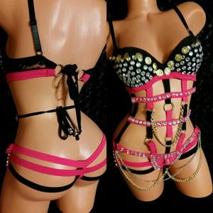May include: A black and pink lingerie set with a bustier top featuring rhinestones and gold coins, and a harness with straps and chains.