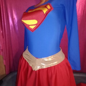 May include: A blue and red superhero costume with a gold emblem. The costume includes a long-sleeved top and a red skirt with a gold belt.