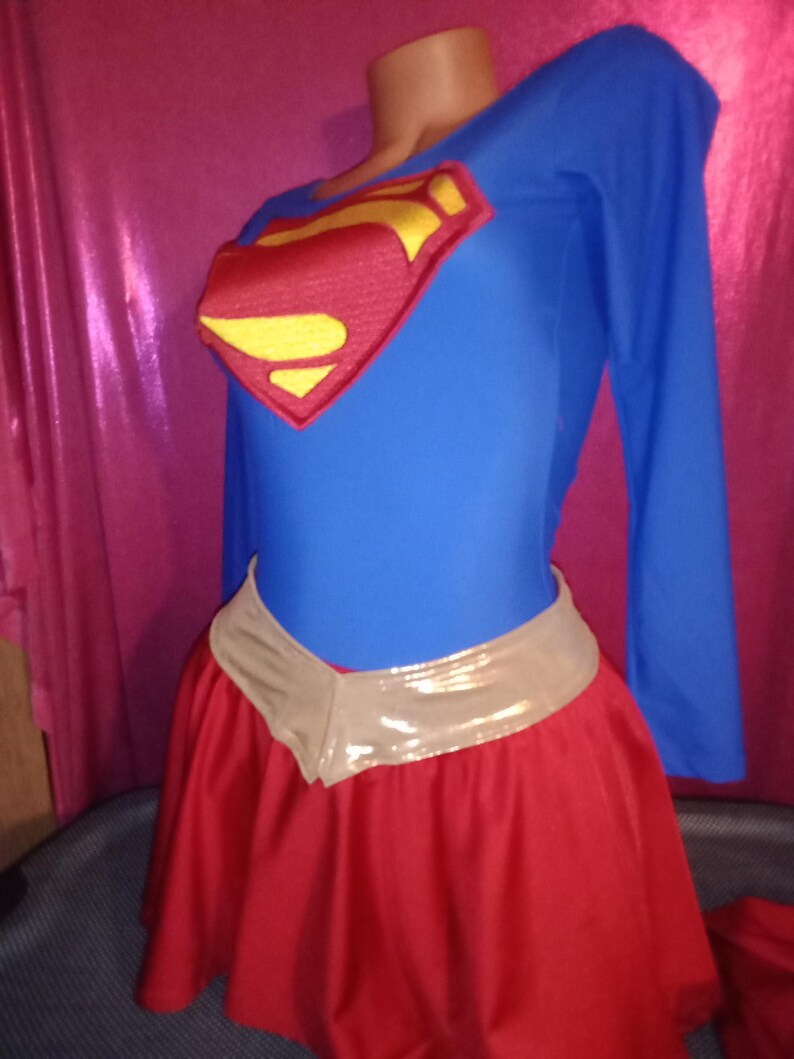 May include: A blue and red superhero costume with a gold emblem. The costume includes a long-sleeved top and a red skirt with a gold belt.