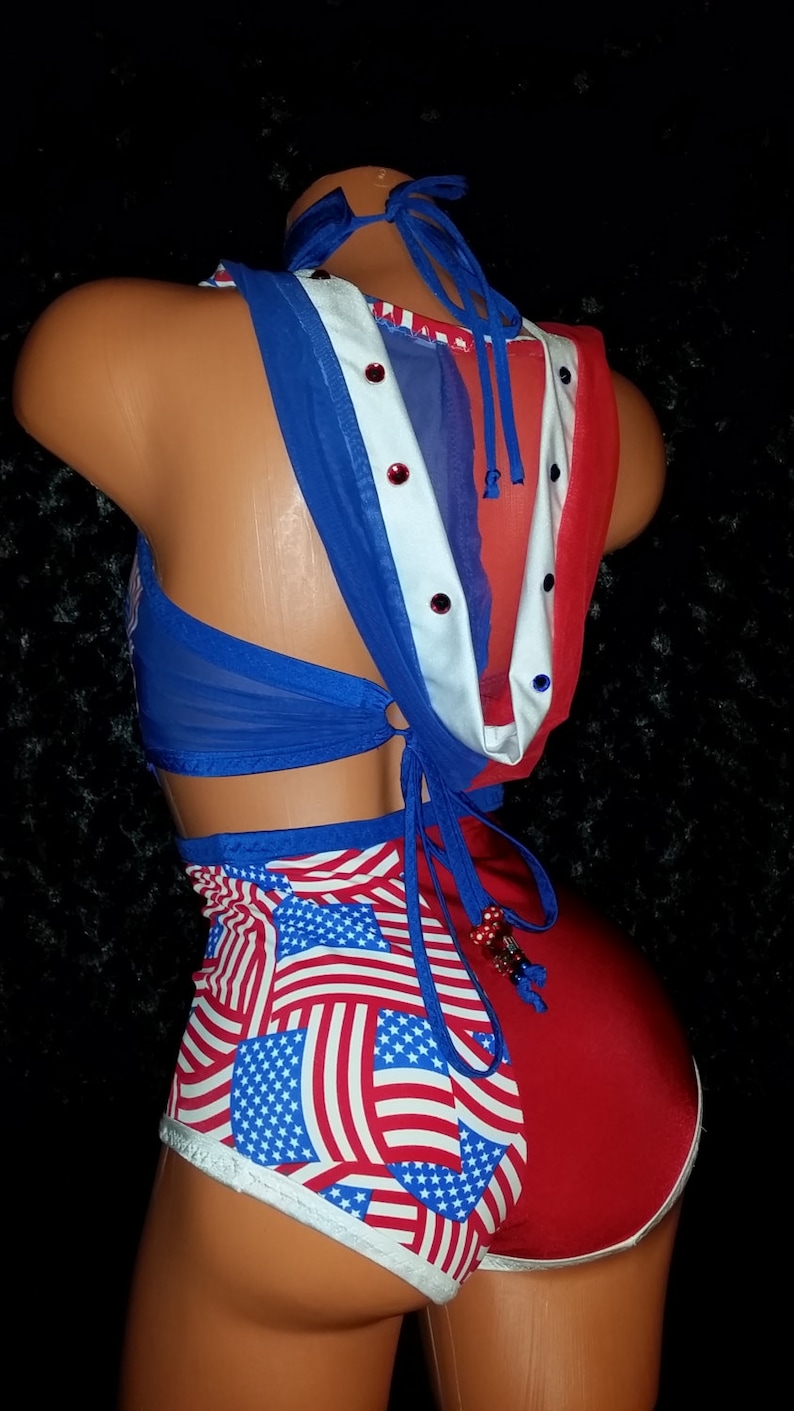 May include: A red, white, and blue two-piece outfit with a mesh top and a bottom featuring a pattern of American flags. The top has a blue tie at the back and is decorated with rhinestones.