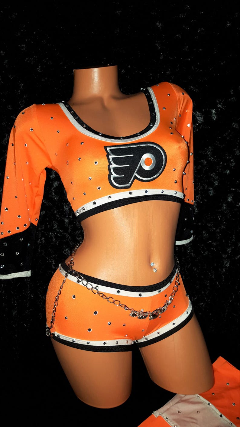May include: Orange and black cheerleading outfit with a white and black trim. The outfit features a crop top with the Philadelphia Flyers logo and a matching skirt with a silver chain.