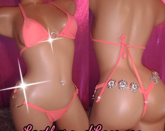 Può includere: Bikini color corallo con top a triangolo e slip a perizoma. Il top presenta un dettaglio con anello metallico e lo slip è impreziosito da strass. Il sito web LeatherandLacz.com è stampato in basso.