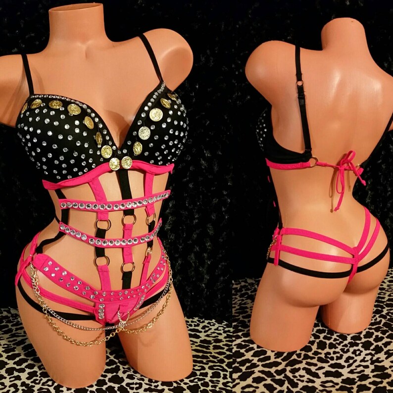 May include: A black and pink lingerie set with a bustier top featuring gold coin accents and rhinestones. The bottom piece is a pink and black harness with a chain detail.