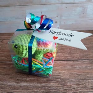 May include: A clear plastic gift box containing a small green crocheted turtle toy resting on colorful paper shreds. The box is tied with a metallic blue ribbon and a white tag with the text Handmade with love and a red heart icon.