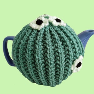 May include: A teal knitted teapot cosy with white and black crocheted flowers on top. The cosy is on a blue teapot.
