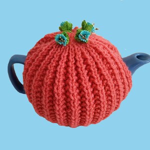 May include: A coral pink knitted teapot cosy with blue and green flower embellishments.