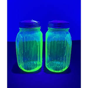 May include: Two clear glass jars with vertical ribbed textures and purple metal lids. Under ultraviolet light, the glass emits a bright neon green glow, highlighting the cylindrical shape and the distinct vertical ridges on the exterior surface.