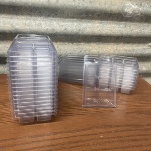 May include: A stack of clear plastic clamshell containers, ideal for packaging small items. The containers are transparent, allowing visibility of the contents. Several other containers are scattered nearby on a wooden surface.