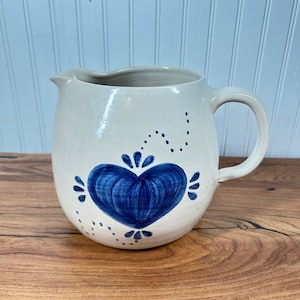 Puede incluir: Una jarra de cerámica de color crema con un diseño de corazón azul pintado a mano en la parte frontal. La jarra tiene un cuerpo redondeado, un asa curva en el lado derecho y un pequeño pico vertedor. El corazón azul tiene pequeños puntos.