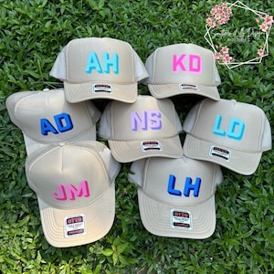 May include: Six beige trucker hats with different colored letters spelling out the names "AH", "KD", "AD", "NS", "LD", and "JM".