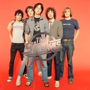 May include: Four individuals standing against a solid orange background. The group wears casual clothing including t-shirts, a button-down shirt, and a blazer. A large, metallic, circular logo with the text The Strokes is overlaid in the center.