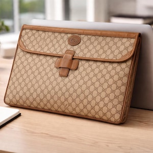 May include: A beige laptop case with a repeating pattern and brown leather trim. The case has a flap closure with a leather buckle. The case is resting against a laptop on a wooden desk, with a notebook and pen in the foreground.