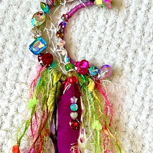 May include: A crescent moon-shaped dream catcher with a purple wrapped frame, adorned with colorful rhinestones and beads. A purple feather is accented with iridescent gems. Strands of green, pink, and purple fabric and yarn hang below.