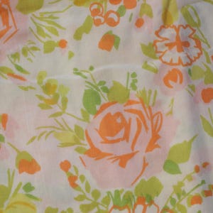 May include: Close-up of a white fabric with a floral pattern. The design features orange, pink, and yellow roses, along with green leaves and smaller orange buds. The flowers are scattered across the fabric.