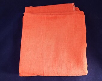 Op de afbeelding: Gevouwen oranje stof, mogelijk linnen of een vergelijkbaar geweven materiaal, tegen een donkerblauwe achtergrond. De stof heeft een getextureerd uiterlijk, met zichtbare draden en een enigszins ongelijkmatig oppervlak. De kleur is een levendig, warm oranje.